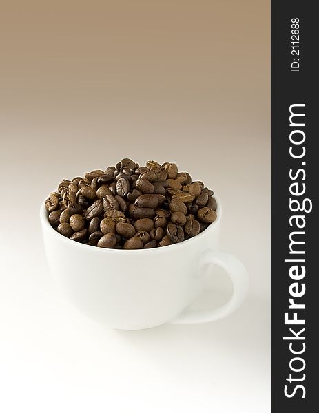 White coffee cup filled with coffee beans on white-brown gradient background. White coffee cup filled with coffee beans on white-brown gradient background.