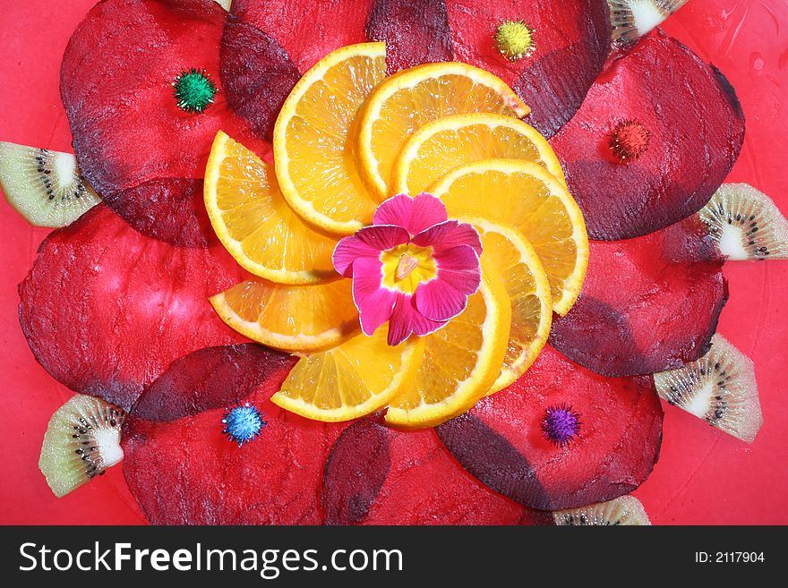 Arrangement of slices of beetroot and orange,with a flower in the middle and pieces of Kiwi,on a glass-plate with a red rim