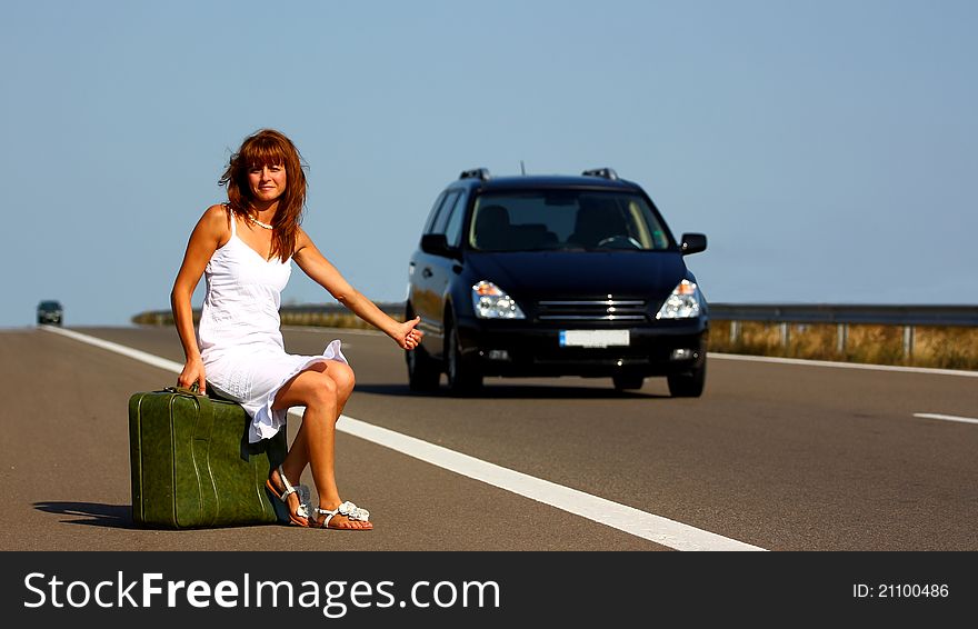 Woman hitchhiking on a highway sitting on a green briefcase