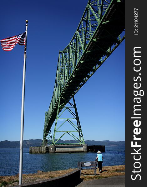 A lady stands under and looks at the historic 4.2 mile long Astoria Megler Bridge which goes over the mouth of the Columbia River in the Pacific Northwest, viewing from the Astoria Oregon side with an American Flag flying. Clear sunny blue skies. 2000 cargo ships a year pass underneath it. A lady stands under and looks at the historic 4.2 mile long Astoria Megler Bridge which goes over the mouth of the Columbia River in the Pacific Northwest, viewing from the Astoria Oregon side with an American Flag flying. Clear sunny blue skies. 2000 cargo ships a year pass underneath it.