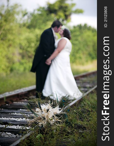 A feather wedding bouquet on railroad tracks with wedding couple standing holding hands behind and out of focus. A feather wedding bouquet on railroad tracks with wedding couple standing holding hands behind and out of focus.