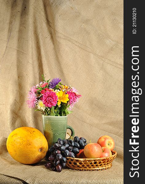 The autumn topic. In the foreground is bouquet of wild and cultivated flowers, bunch of black grapes and melon. They are on the table covered by rough stuff. This staff creates background. The autumn topic. In the foreground is bouquet of wild and cultivated flowers, bunch of black grapes and melon. They are on the table covered by rough stuff. This staff creates background.