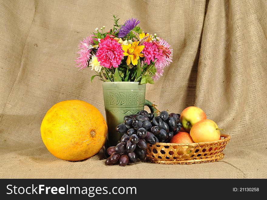 The autumn topic. In the foreground is bouquet of wild and cultivated flowers, bunch of black grapes and melon. They are on the table covered by rough stuff. This staff creates background. The autumn topic. In the foreground is bouquet of wild and cultivated flowers, bunch of black grapes and melon. They are on the table covered by rough stuff. This staff creates background.