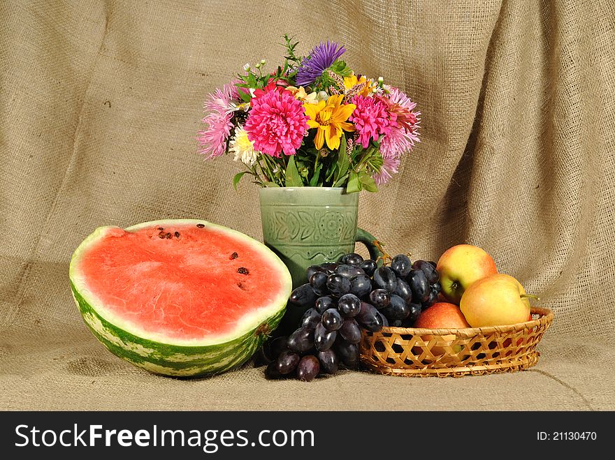 The autumn topic. In the foreground is bouquet of wild and cultivated flowers, bunch of black grapes and melon. They are on the table covered by rough stuff. This staff creates background. The autumn topic. In the foreground is bouquet of wild and cultivated flowers, bunch of black grapes and melon. They are on the table covered by rough stuff. This staff creates background.