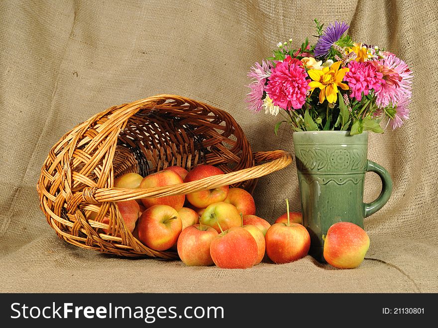 The autumn topic. In the foreground is bouquet of wild and cultivated flowers and basket with red apples. The basket lies sideways and apples scatter over the table. The table is covered by rough stuff. This staff creates background. The autumn topic. In the foreground is bouquet of wild and cultivated flowers and basket with red apples. The basket lies sideways and apples scatter over the table. The table is covered by rough stuff. This staff creates background.