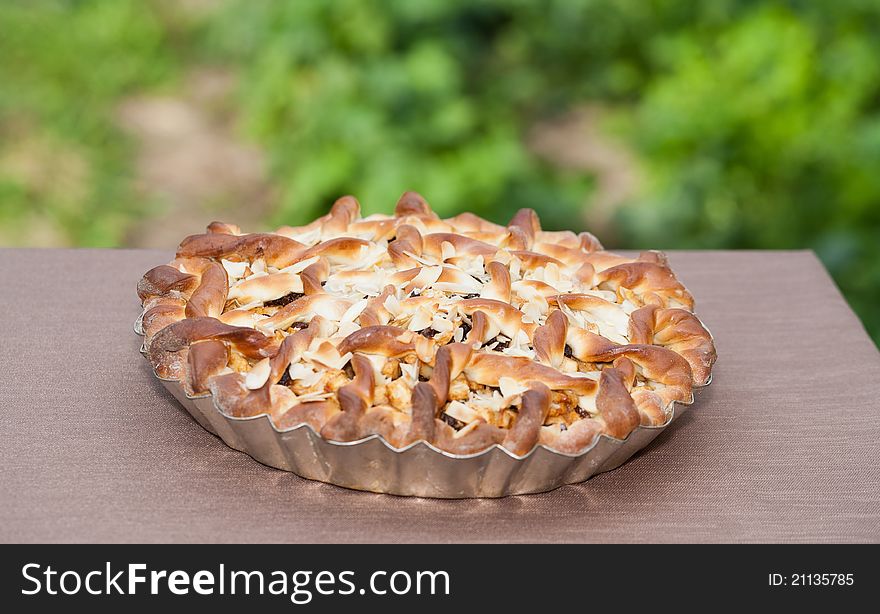 An outdoors angled closeup of a freshly baked homemade Belgian apple-raisin pie topped with almond slices sitting on a tablecloth with the garden in the background. An outdoors angled closeup of a freshly baked homemade Belgian apple-raisin pie topped with almond slices sitting on a tablecloth with the garden in the background.