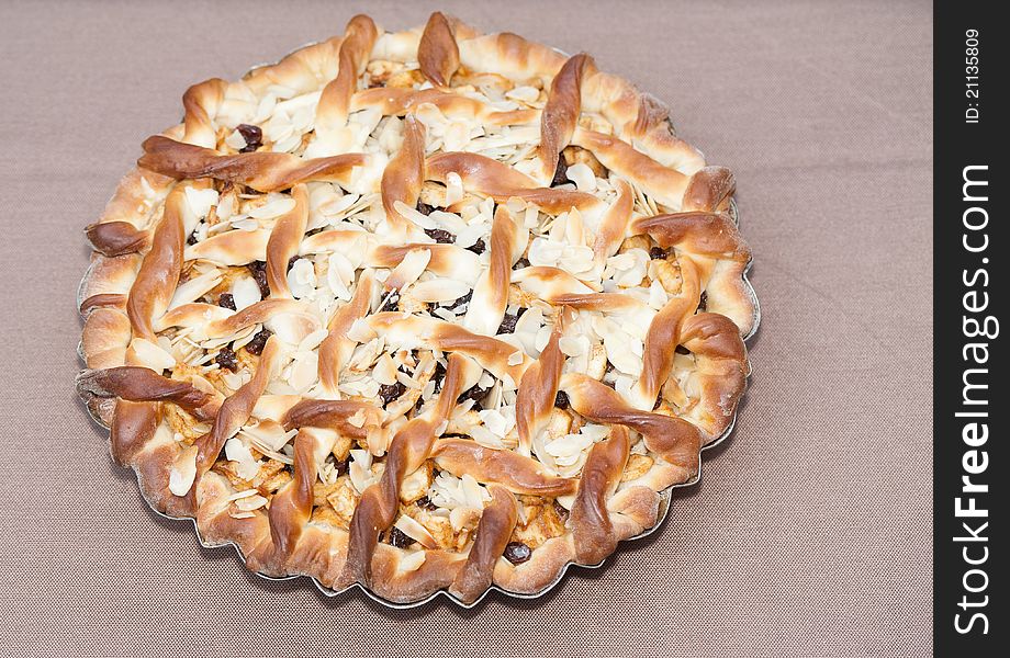 An outdoors top closeup of a homemade Belgian apple-raisin pie topped with almond slices sitting on a tablecloth. An outdoors top closeup of a homemade Belgian apple-raisin pie topped with almond slices sitting on a tablecloth.