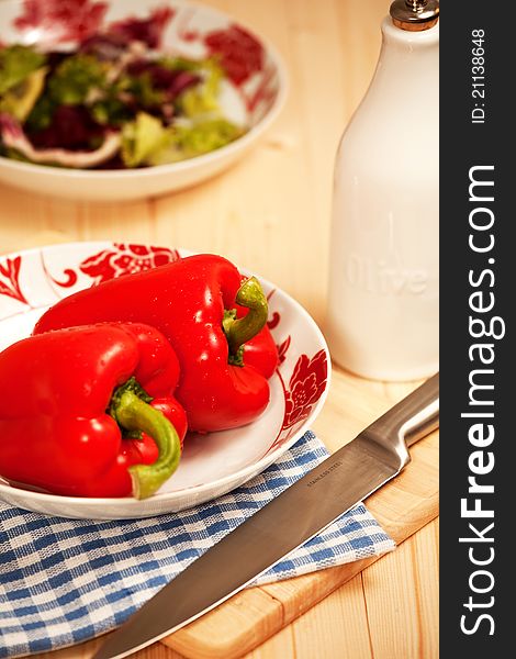 Two red peppers on a table with silver knife and salad in the background.