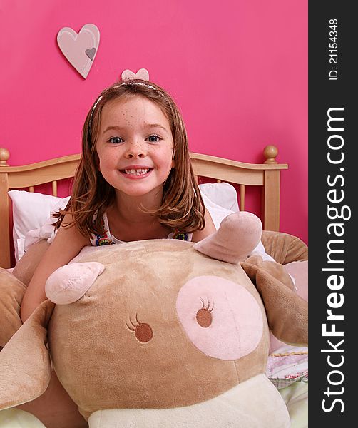 A really cute young girl playing with a giant teddy in a girly pink room whilst sat on a bed.
