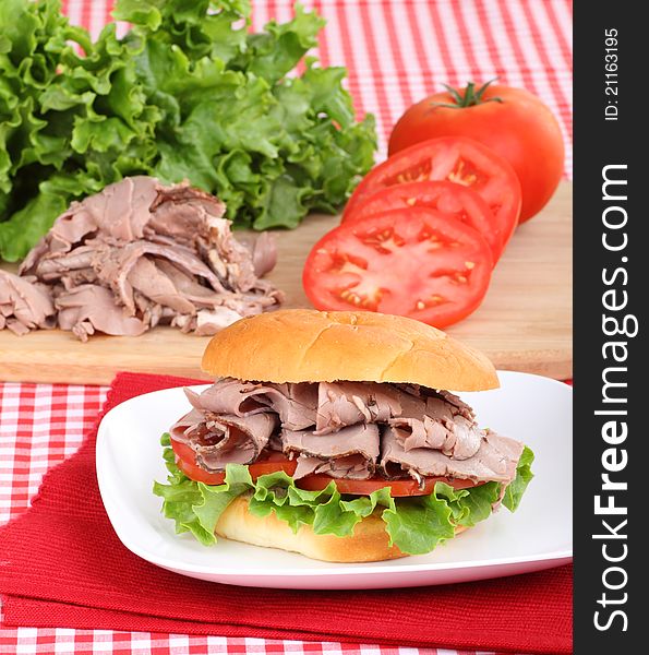 Roast beef sandwich with lettuce and tomato on a plate with ingredients in background. Roast beef sandwich with lettuce and tomato on a plate with ingredients in background