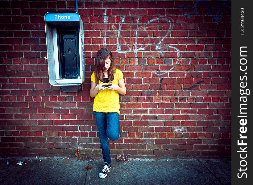 A cute teenage girl standing next to an obsolete payphone while texting on a cell phone against a grungy urban wall. A cute teenage girl standing next to an obsolete payphone while texting on a cell phone against a grungy urban wall