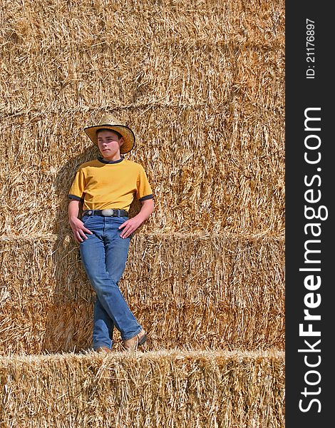 Male cowboy standing on straw stack with cowboy hat wearing yellow shirt and blue jeans in the sun looking off in the distance. Male cowboy standing on straw stack with cowboy hat wearing yellow shirt and blue jeans in the sun looking off in the distance