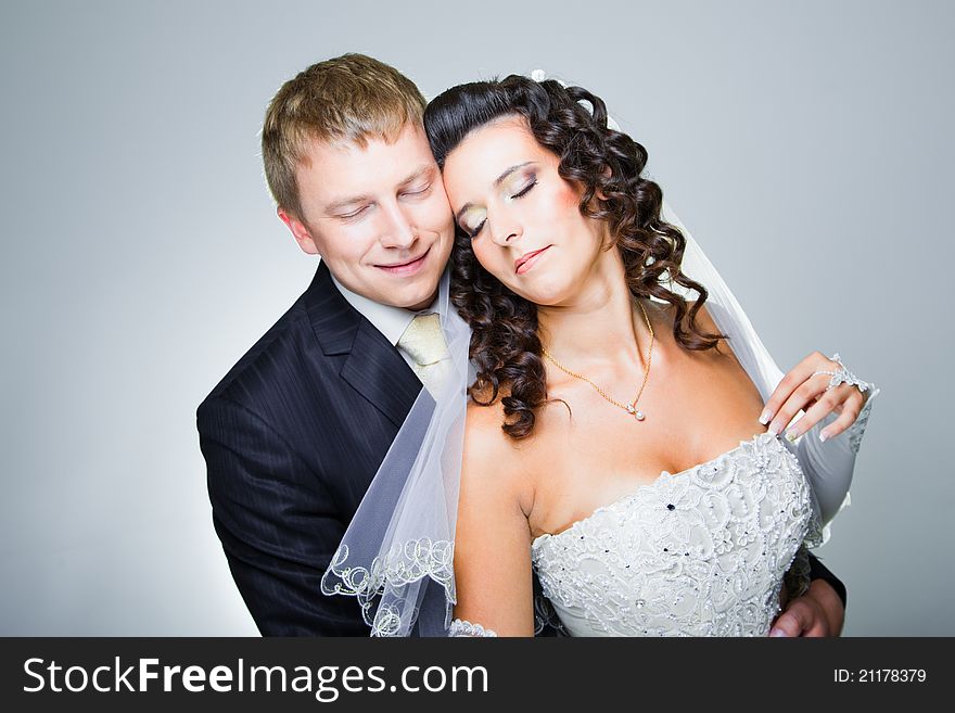 Studio portrait of young beautiful happy just married bride and groom with closed eyes embracing on blue background