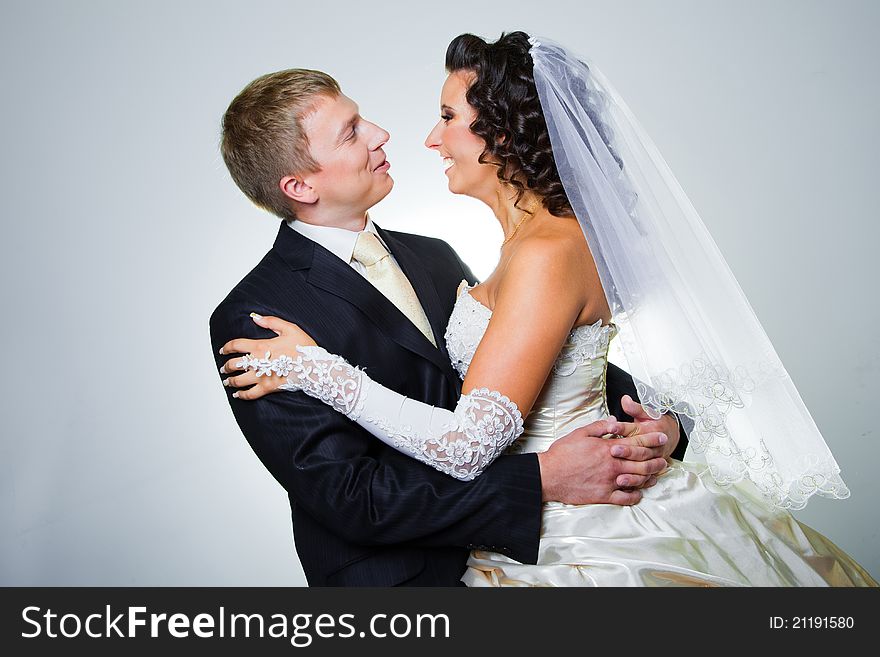 Studio portrait of young elegant enamored just married bride and groom looking to each other and embracing on grey background