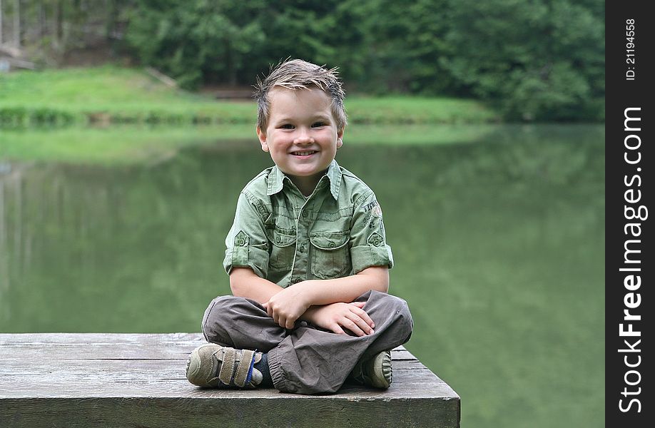 Picture of a young caucasian white boy sitting cross-legged on a bench at a pond or lake smiling. Picture of a young caucasian white boy sitting cross-legged on a bench at a pond or lake smiling