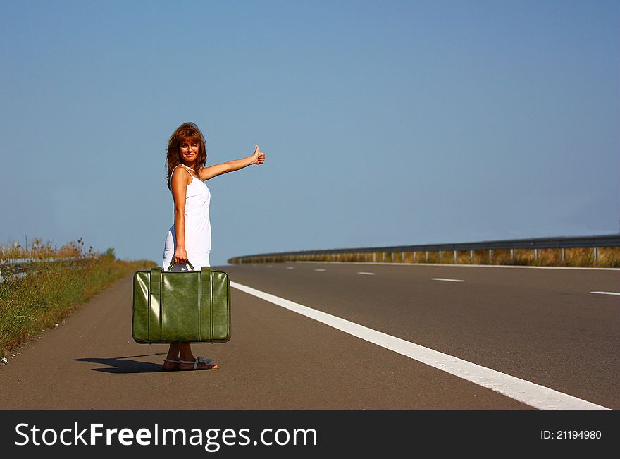 Woman hitchhiking on a empty highway holding a green briefcase on a clear blue sky background