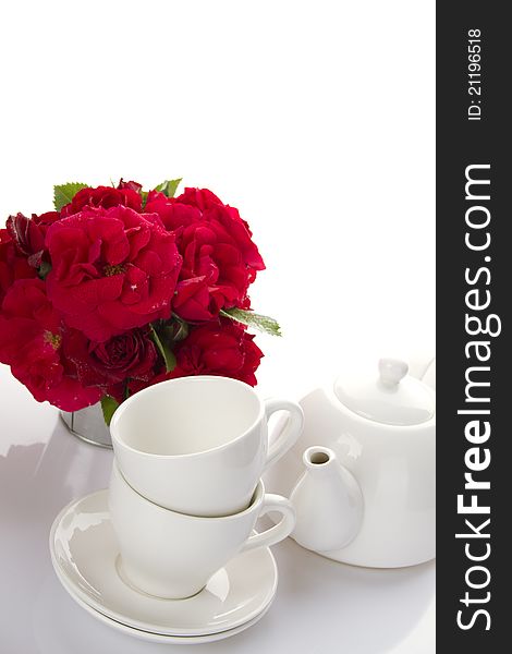 White crockery for tea kettle, two cups and saucers and a bouquet of red roses in a small iron bucket. White crockery for tea kettle, two cups and saucers and a bouquet of red roses in a small iron bucket