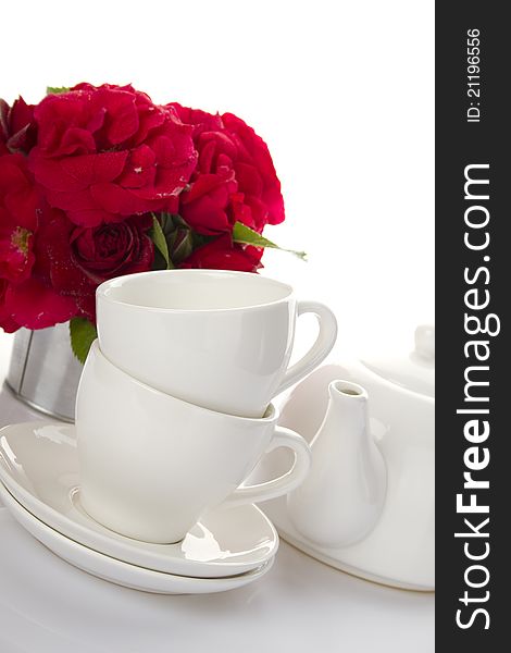 White crockery for tea kettle, two cups and saucers and a bouquet of red roses in a small iron bucket. White crockery for tea kettle, two cups and saucers and a bouquet of red roses in a small iron bucket