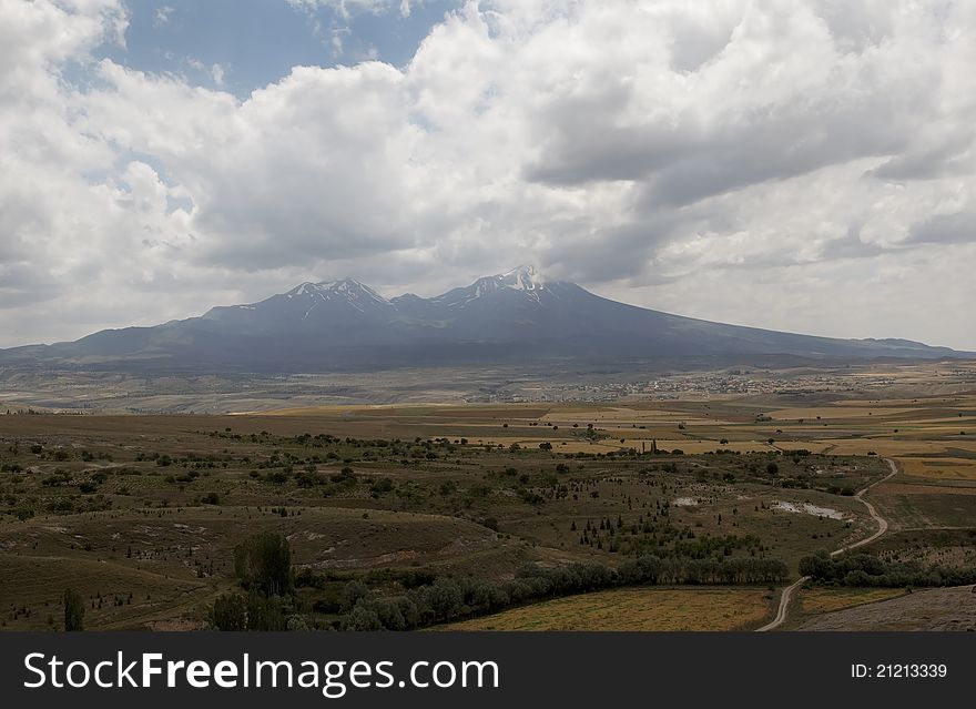 Long wind dirt track over plains to the snow capped mountains of Cappadocia, Turkey. Horizontal landscape under cloudy blue sky with crop space and copy area. Long wind dirt track over plains to the snow capped mountains of Cappadocia, Turkey. Horizontal landscape under cloudy blue sky with crop space and copy area