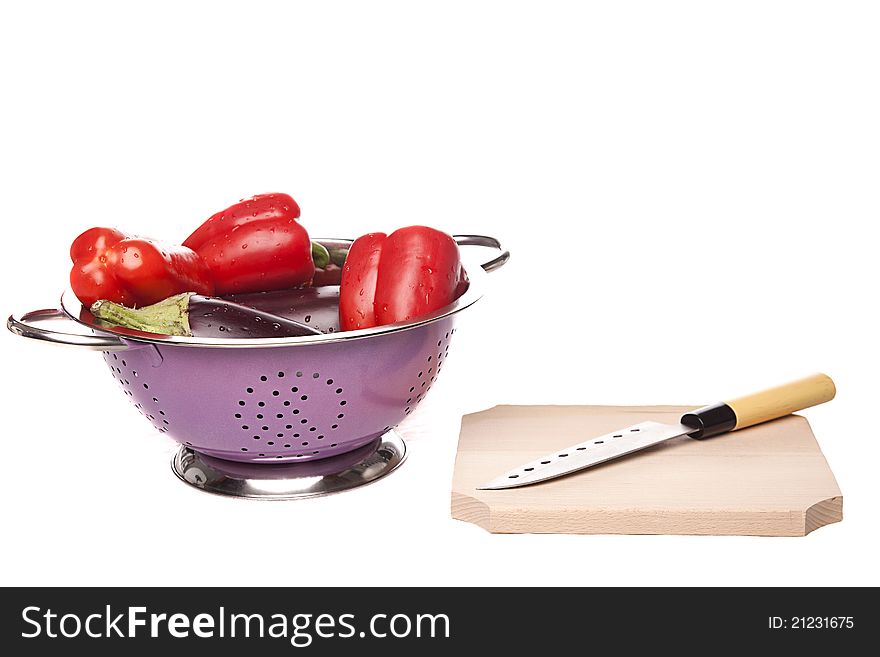 Red peppers and eggplants in a pink bowl next to a wood cutting board and a sharp knife on a white background. Red peppers and eggplants in a pink bowl next to a wood cutting board and a sharp knife on a white background.