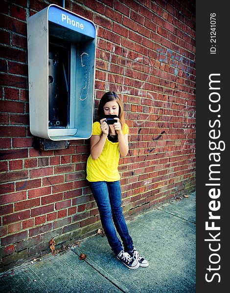 A cute teenage girl texting on her cell phone against a grungy urban wall with an obsolete payphone. A cute teenage girl texting on her cell phone against a grungy urban wall with an obsolete payphone