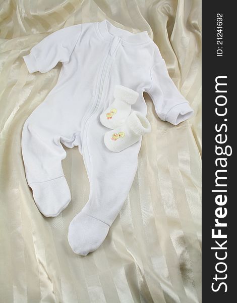 White baby romper suit on cream satin background. White booties with yellow embroidered chicks. White baby romper suit on cream satin background. White booties with yellow embroidered chicks.