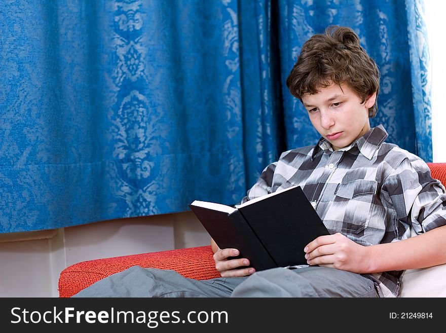 A pre-teen boy in a checkered shirt reading a blank black book on an orange couch with blue damask curtains in the background. A pre-teen boy in a checkered shirt reading a blank black book on an orange couch with blue damask curtains in the background.