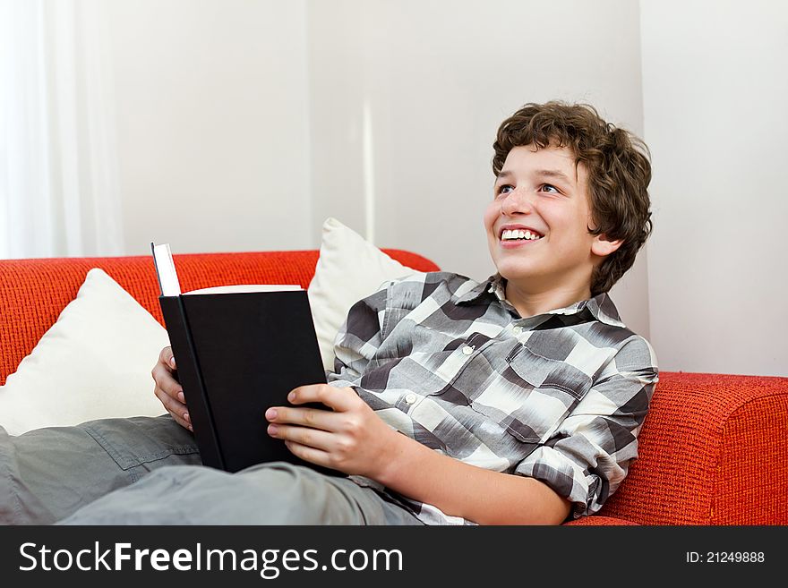 A preteen boy laying back on an orange couch with white pillows as he holds a black book in his hand and looks up with a big smile. A preteen boy laying back on an orange couch with white pillows as he holds a black book in his hand and looks up with a big smile.