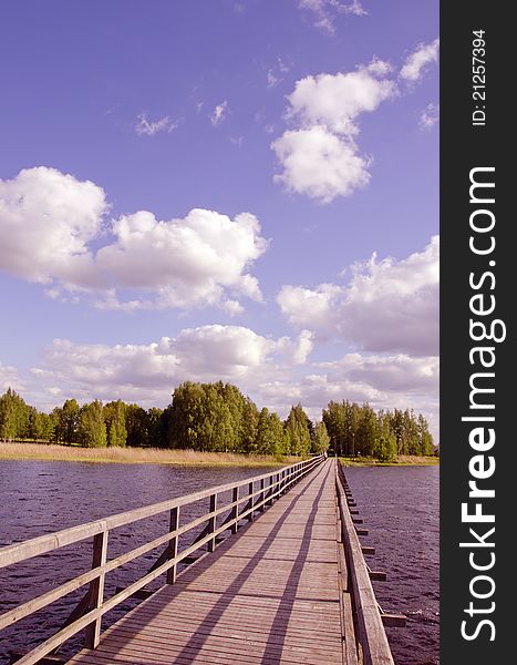 Long wooden footbridge with handrails over the lake and the man walking far away. Forest in the distance and the cloudy sky. Long wooden footbridge with handrails over the lake and the man walking far away. Forest in the distance and the cloudy sky.