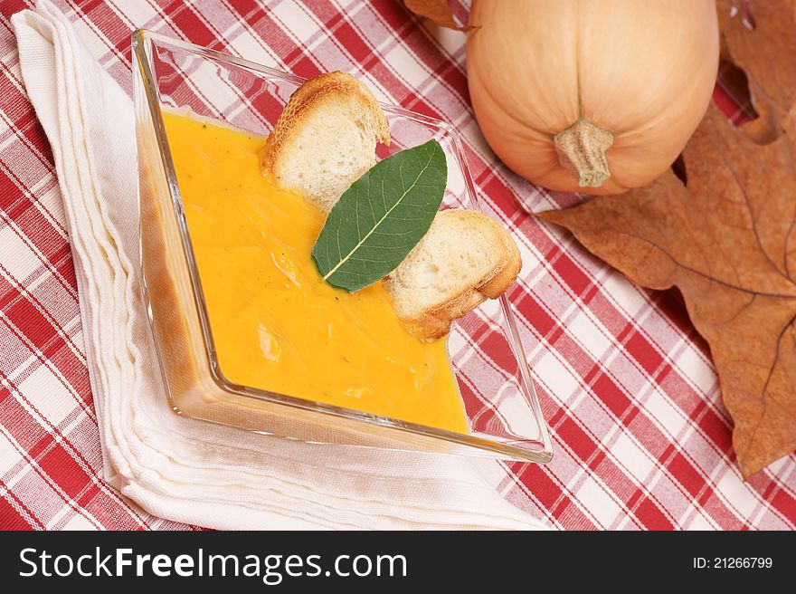 Cream of squash soup in a glass plate with toasted bread and leaf bay. A whole pumpkin and autumn leaves are out of focus in the background. Selective focus, shallow DOF.