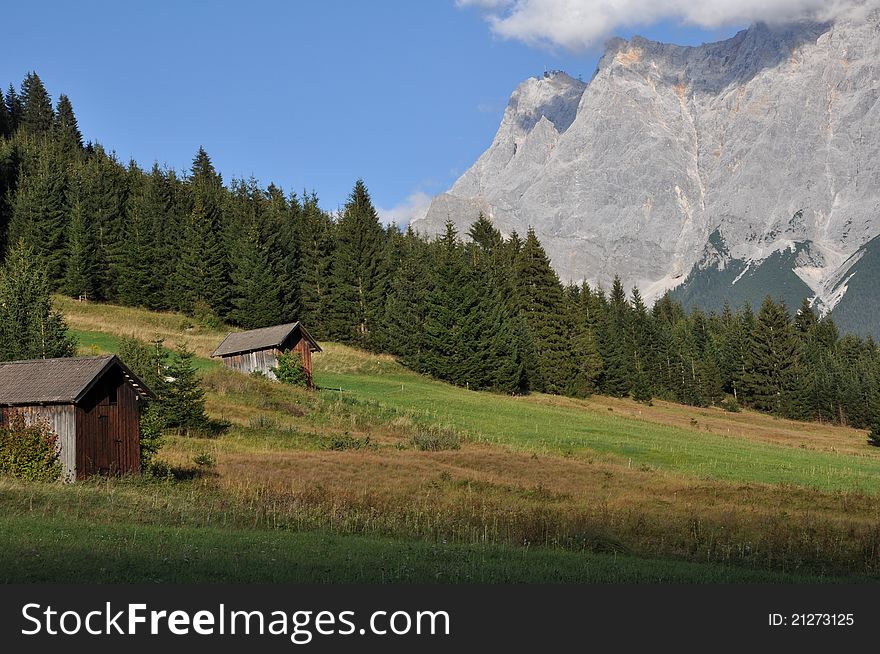 Image shows the highest mountain of Germany called Zugspitze on the right with blue sky and some clouds. In the front there is a green and brown meadow and some green pines. On the left are two wooden houses. Image shows the highest mountain of Germany called Zugspitze on the right with blue sky and some clouds. In the front there is a green and brown meadow and some green pines. On the left are two wooden houses.