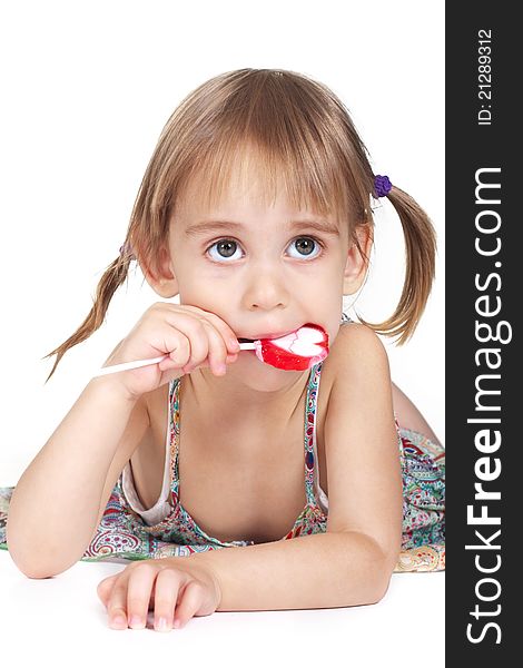 Little girl with big bright eyes lying on front and eating red heart-shaped lollipop on white background studio shot. Little girl with big bright eyes lying on front and eating red heart-shaped lollipop on white background studio shot