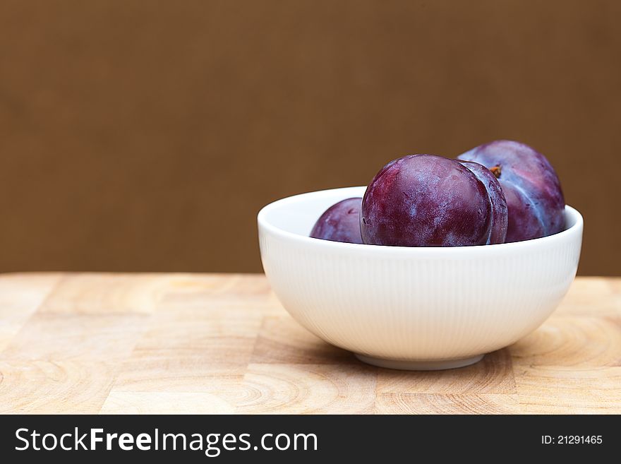 Some plumbs in a simple white bowl on a wooden table.  Focus on the front plumb.  Plain brown background. Some plumbs in a simple white bowl on a wooden table.  Focus on the front plumb.  Plain brown background.