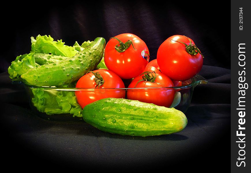 Tomatoes, cucumbers, and leaves of salad in a roaster, on a black background with soft illumination. Tomatoes, cucumbers, and leaves of salad in a roaster, on a black background with soft illumination