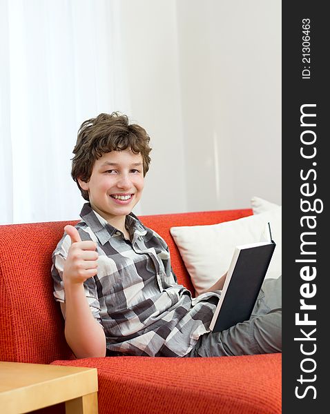 A preteen boy seated on an orange couch with white pillows gives a thumbs up and a big smile as he holds the book he was reading with his other hand. A preteen boy seated on an orange couch with white pillows gives a thumbs up and a big smile as he holds the book he was reading with his other hand.