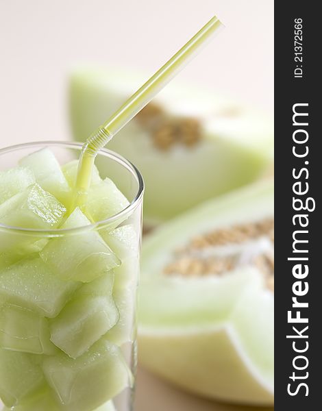 Green honeydew melon pieces in a glass with straw and blurred melon in background. Green honeydew melon pieces in a glass with straw and blurred melon in background