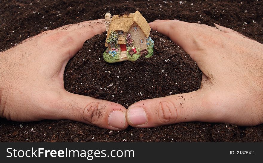 Two hands on brown soil making a circle with small country house in center. Two hands on brown soil making a circle with small country house in center