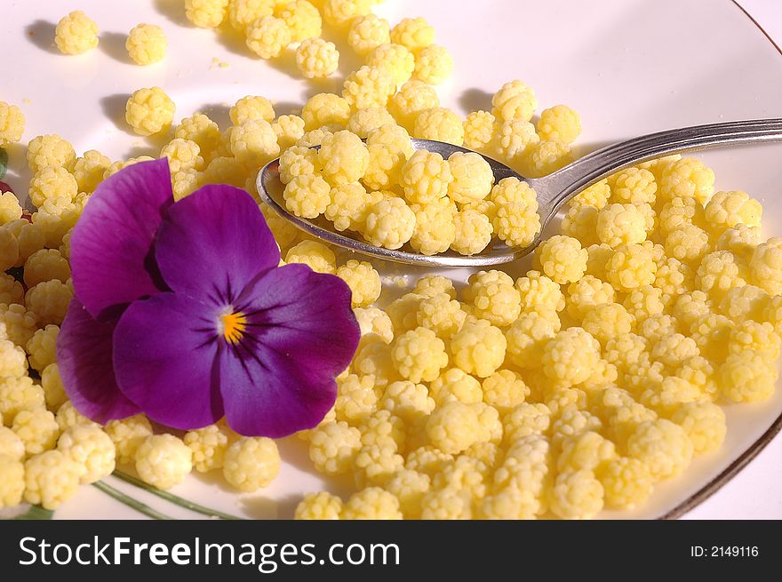 Small yellow candies like pollen with purple pansy an teaspoon on a saucer