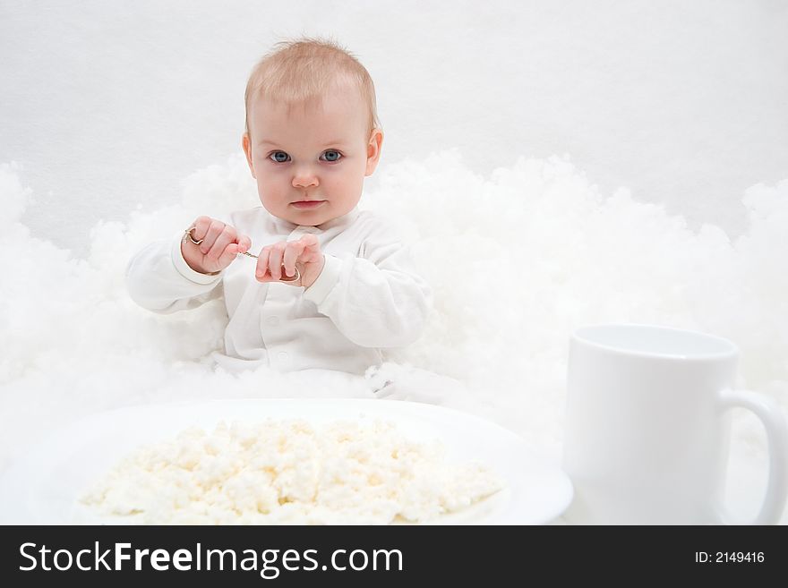 Cute little girl sitting on white blanket with spoon in her hands. There are white plate with curd and white cup in front of her. Soft-focused, focal point is on the girl. Cute little girl sitting on white blanket with spoon in her hands. There are white plate with curd and white cup in front of her. Soft-focused, focal point is on the girl