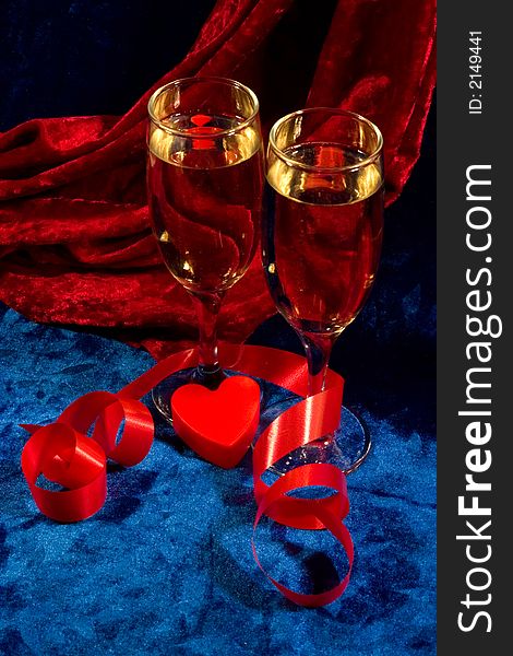 Two glasses of champagne, red twisted ribbon, and plastic heart on the blue velvet