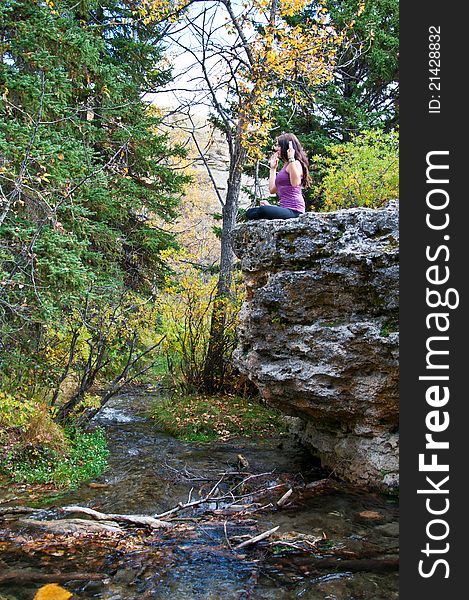 Young attractive girl sitting on the edge of a rock in autumn doing yoga and meditating with a creek running beneath her. Young attractive girl sitting on the edge of a rock in autumn doing yoga and meditating with a creek running beneath her