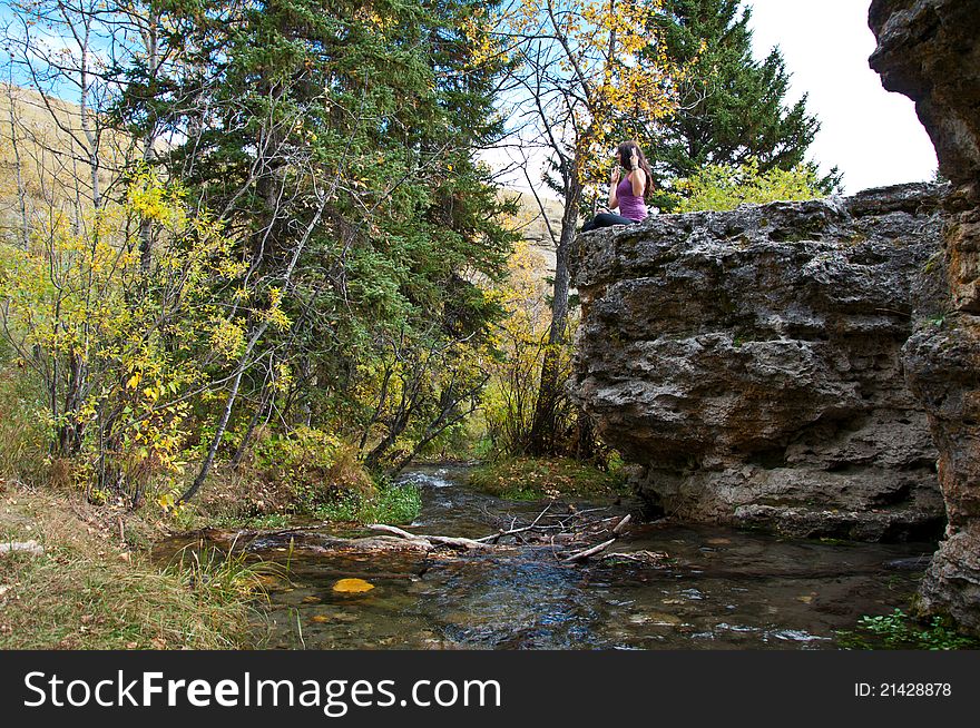 Young attractive girl sitting on the edge of a rock in autumn doing yoga and meditating with a creek running beneath her. Young attractive girl sitting on the edge of a rock in autumn doing yoga and meditating with a creek running beneath her