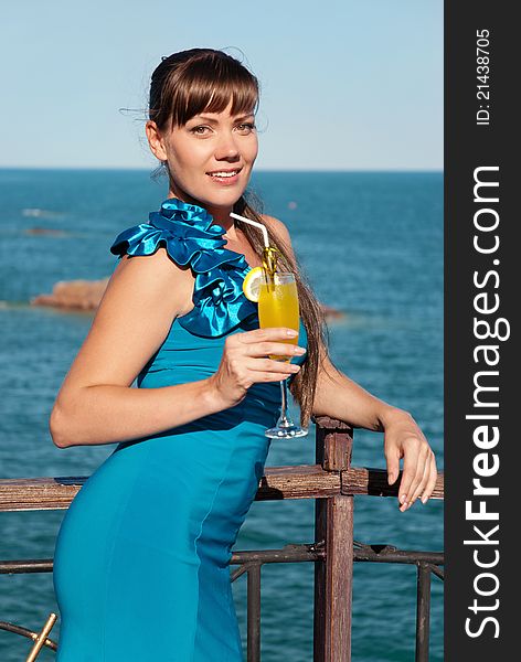 Beautiful young girl in a smart blue dress stands against the sea. She smiles and looks pretty happy. The girl in the hand of a bright yellow drink or juice. Beautiful young girl in a smart blue dress stands against the sea. She smiles and looks pretty happy. The girl in the hand of a bright yellow drink or juice.