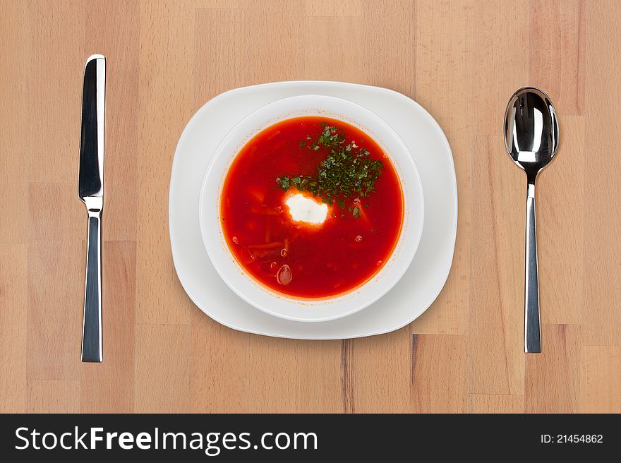 A plate of traditional Russian red borsch with sour cream and greens on a wooden table with spoon and knife.