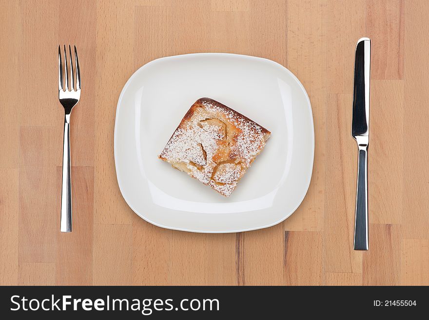 A plate with piece of baked pudding powdered with sugar with fork and knife on a wooden table.