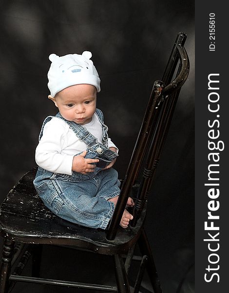 Image of adorable baby wearing denim overalls sitting on a black chair. Image of adorable baby wearing denim overalls sitting on a black chair