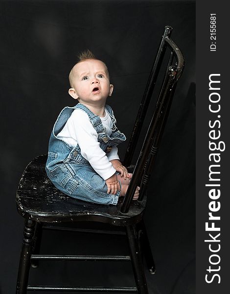 Image of adorable baby wearing denim overalls sitting on a black chair. Image of adorable baby wearing denim overalls sitting on a black chair