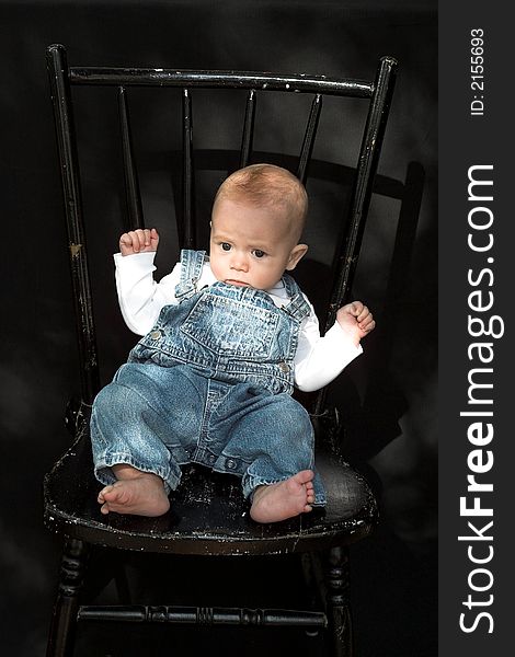 Image of adorable baby wearing denim overalls sitting on a black chair. Image of adorable baby wearing denim overalls sitting on a black chair