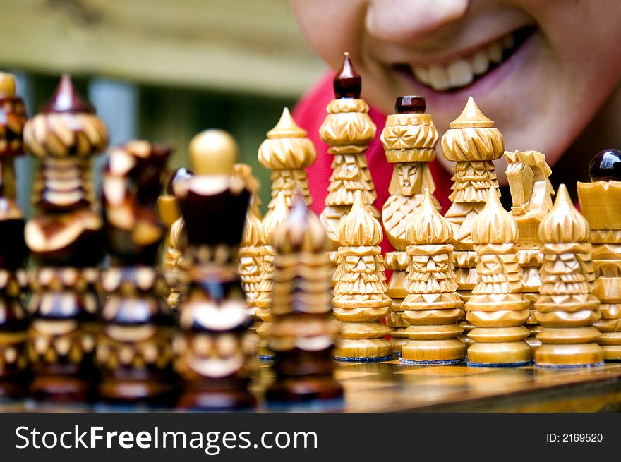 A boy's smiling mouth is visible behind the white pieces as he leans over an ornate wooden chess set assembled for the beginning of a match. A boy's smiling mouth is visible behind the white pieces as he leans over an ornate wooden chess set assembled for the beginning of a match.