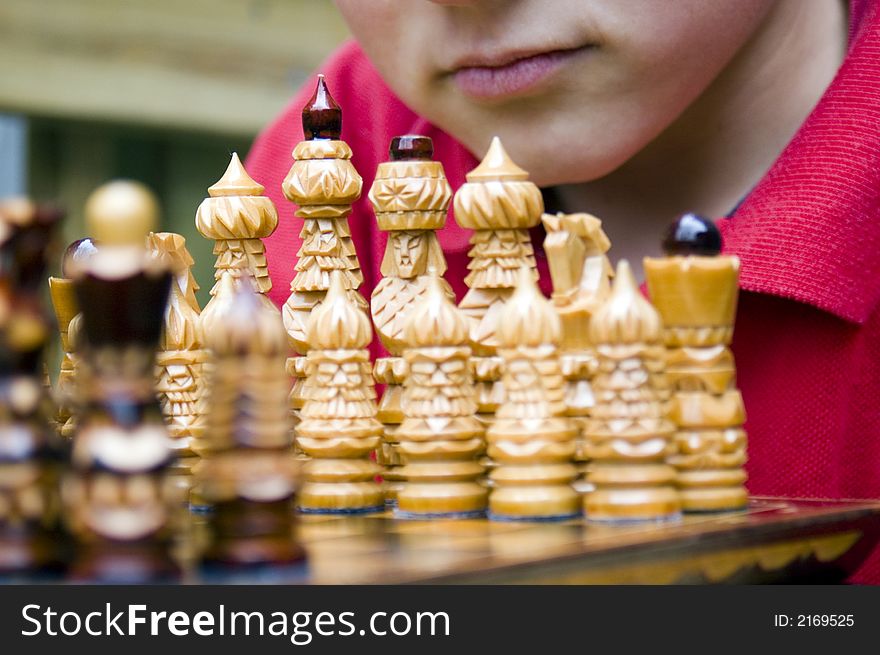 A boy's mouth with serious expression is visible behind the white pieces as he leans over an ornate wooden chess set assembled for the beginning of a match. A boy's mouth with serious expression is visible behind the white pieces as he leans over an ornate wooden chess set assembled for the beginning of a match.